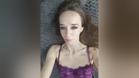 Video of a pale-skinned woman with long brown hair, wearing a purple lace-trimmed camisole, lying on a grey geometric-patterned blanket, with a blurred, partially visible person in the foreground.