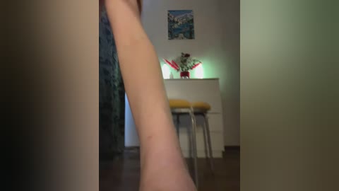 Video of a person's arm extending through a doorway into a dimly lit room with a white desk, yellow chair, and colorful flower arrangement.