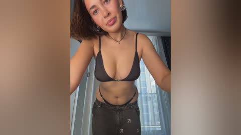 A video of a young woman with medium skin tone and short brown hair, wearing a black bra and high-waisted pants, standing indoors with a neutral expression.