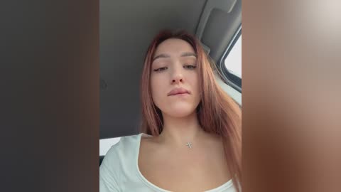 Video of a young woman with long, straight auburn hair, fair skin, and light makeup, wearing a white top. She has a subtle, confident expression. Background includes a car window and a cross necklace.