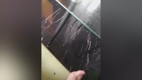 A video of a person's hand with a purple nail polish touching a dark, wet surface with white streaks, likely from rain or water, suggesting a rainy outdoor scene.