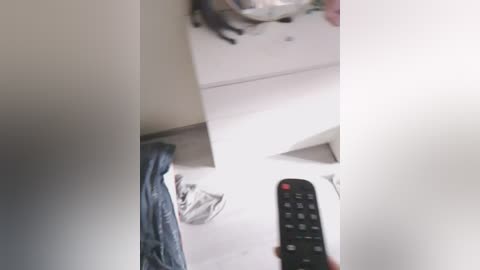 A video shows a person holding a black remote control in front of a white tiled wall. The person's hand is partially visible, and a gray jacket is draped over the wall.