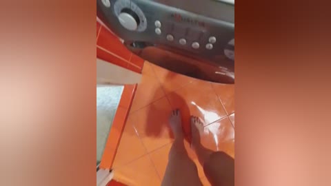 Video of a person's foot in an orange-tiled bathroom, with a grey control panel above. The image is slightly blurred, possibly due to movement.