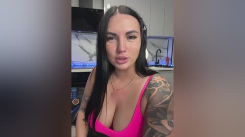 Video of a tattooed, light-skinned woman with long black hair, wearing a low-cut pink tank top, sitting in a modern kitchen with marble countertops and white cabinets.