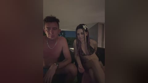 Video: A shirtless, dark-haired man with a silver chain sits next to a dark-haired woman in a bikini, smiling. They stand in a dimly lit room with blurred furniture in the background.