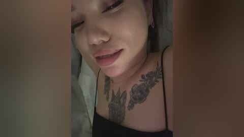 A close-up video of a young woman with light skin, dark hair, and a black tank top, showing a detailed floral tattoo on her chest. She has a relaxed expression with her eyes closed.