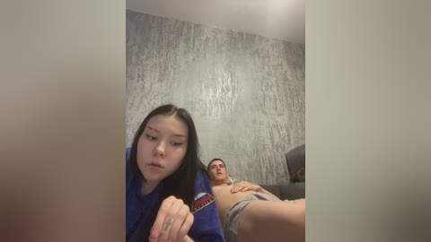 A video of an Asian woman with long black hair, wearing a blue shirt, and a Caucasian man with short dark hair, lying on a bed with gray sheets. The background features a textured gray wall.