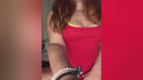 A video of a fair-skinned woman with long, wavy auburn hair, wearing a tight, red strapless top that reveals a yellow bra. She has a white beaded bracelet on her right wrist. The background is a plain, light-colored wall with a red vertical stripe.