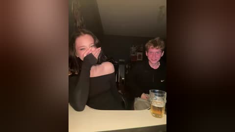 Video of a young woman with dark hair, wearing a black dress, sitting at a table, hiding her face with her hands. Behind her, a young man with curly hair, wearing glasses and a black hoodie, looks on.