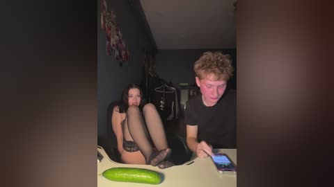 Video of a woman in black lingerie, sitting on a table with a cucumber, next to a man in a black t-shirt, holding a phone.
