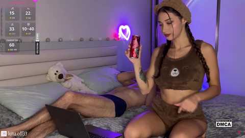 Video of a woman in a bear costume, holding a can, sitting on a bed with a man and a stuffed bear.