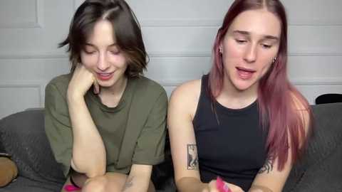 Video of two women with short hair, one with olive skin and brown hair, the other with pale skin and red hair, sitting on a couch, smiling, wearing casual black tops.