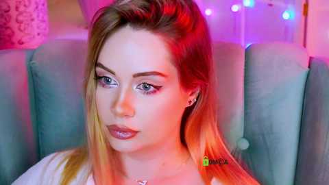 Video of a young woman with fair skin and long blonde hair, wearing black eyeliner and a white top, sitting in a light blue armchair with colorful LED lights in the background.