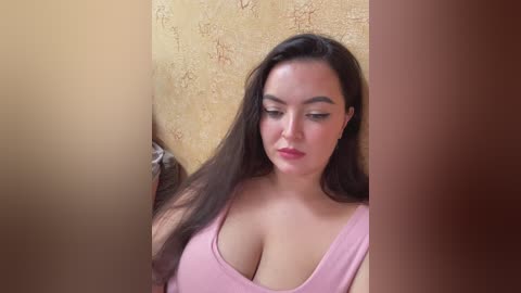 Video of a young woman with long dark hair, wearing a pink tank top, standing in a narrow, dimly lit corridor with yellowish-brown textured walls.