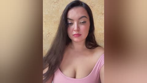 A video of a Latina woman with long dark hair, wearing a pink off-shoulder top, against a textured yellow wall.
