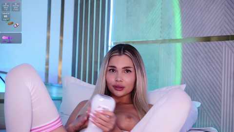 Video of a topless woman with long blonde hair, light skin, and medium-sized breasts, lying on a bed, holding a white phone. The background features a modern, white, and blue room with geometric patterns.