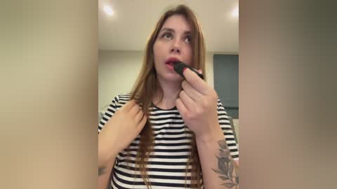 Video of a young woman with long, straight auburn hair, wearing a black-and-white striped shirt, applying black eyeliner with a small brush. She has a tattoo on her left forearm. The background is a neutral-toned room with recessed lighting.