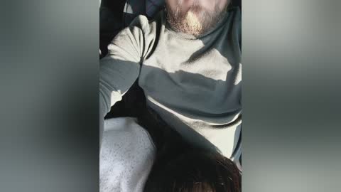 A video captures a man with a full beard and light brown hair, wearing a gray sweater and a white long-sleeved shirt. He's sitting in a car, with shadows casting on his face and clothing.