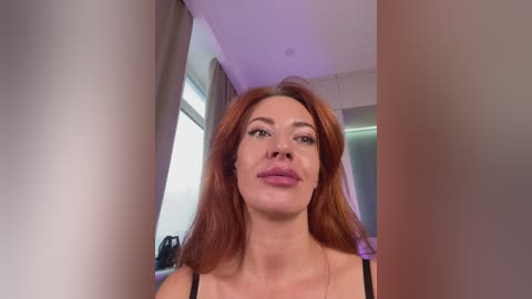 A video of a fair-skinned woman with long, auburn hair, wearing a black tank top, standing indoors with white walls and a window in the background.