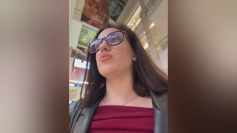 Video of a young woman with long brown hair, wearing black-rimmed glasses, a maroon top, and a black leather jacket, standing indoors in a modern, bright, glass-walled building.
