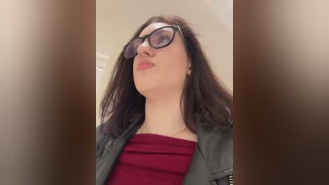 Video of a young woman with long brown hair, wearing black glasses, a red top, and a green jacket, standing indoors with blurred background.