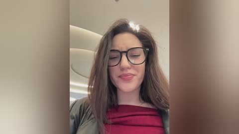 Video of a young woman with fair skin, brown hair, and glasses, wearing a red top, smiling. Background shows white walls and a blurred staircase.