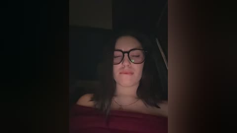 A dimly lit video captures a woman with long dark hair, glasses, and a maroon off-shoulder top, smiling with eyes closed. The background is dark and indistinct.