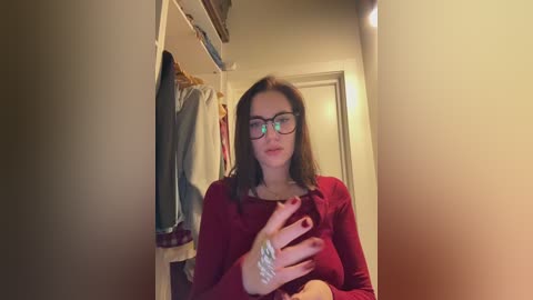 Video of a young woman with long brown hair, wearing black-framed glasses, a red sweater, and a silver bracelet. She is seated in a dimly lit closet with hanging clothes in the background.