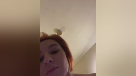 Video of a young woman with fair skin and red hair, eyes closed, lying on a bed with beige walls, wearing minimal makeup.