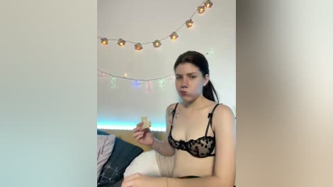 A video of a young woman with pale skin and brown hair, wearing black lace lingerie, sitting on a bed with a festive string of lights and colorful banners in the background.