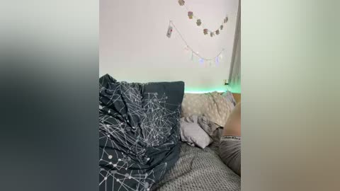 Video of a cozy, cluttered room with a dark gray couch covered in a white starry-patterned blanket, light green string lights, and a messy bed in the background.