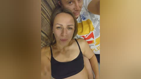 A video of a woman with light skin and straight brown hair, wearing a black bra, smiling beside a man in a yellow and white t-shirt, both sitting on a textured brown couch.