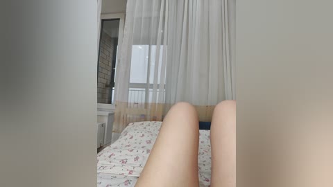 Video of a woman's bare legs on a bed with floral sheets, sheer white curtains, and a brick wall in the background, creating a cozy, intimate bedroom scene.