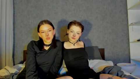 Video of two young women, one with straight hair, black dress, and choker, sitting on a bed with grey bedding, in a dimly lit room with a grey textured wall.