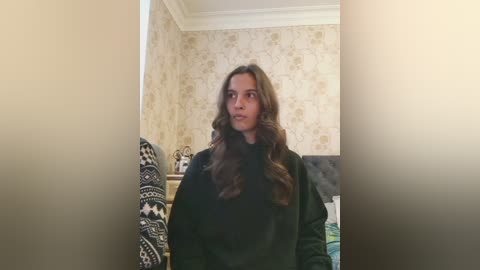 Video of a young woman with long, wavy brown hair, wearing a black jacket, standing in a room with beige floral wallpaper and a bed with a patterned blanket.