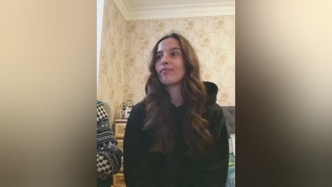 A video of a young woman with wavy, light brown hair, wearing a black hoodie, standing in a dimly lit room with beige wallpaper and a patterned cushion on a wooden chair.