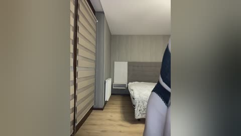 A video of a minimalist bedroom with a white bed, grey wall, and beige striped curtains, seen through a doorway, featuring a person in blue underwear.