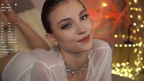 Video of a fair-skinned woman with dark hair, wearing a sheer white top, silver necklace, and a warm smile. Background features bokeh lights and a cozy setting.