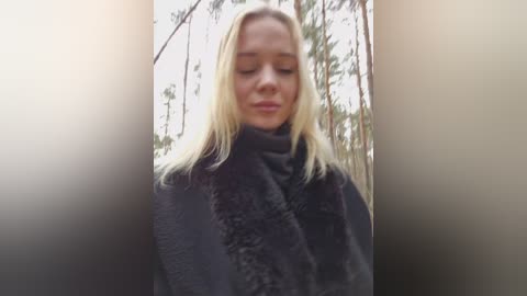 Video of a blonde woman with fair skin, wearing a dark, textured scarf, standing in a forest. The background features tall pine trees and a soft, diffused light. The image has a soft focus and warm lighting.