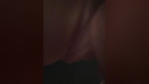 A blurry video showing a close-up of a person's chest and shoulder, with visible skin texture and a black garment partially covering the lower region. The background is a dark, reddish-brown color.