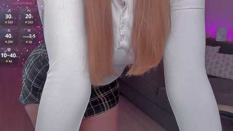 Video of a young woman with long, straight blonde hair, wearing a white long-sleeve top, black plaid skirt, and white thigh-high stockings. Background includes a pink wall and a gray sofa.