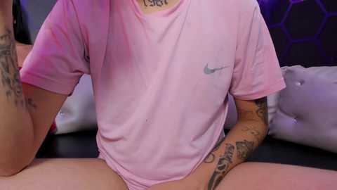 A video of a person with light skin, wearing a pink Nike t-shirt and black underwear, sitting cross-legged on a black couch with purple hexagonal pillows, tattoos on their arms.