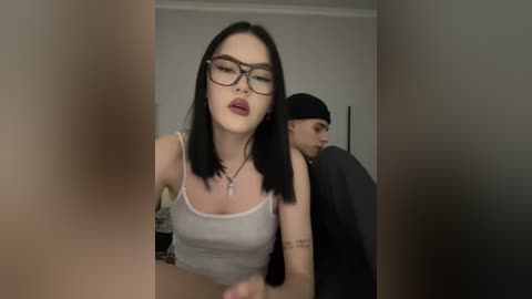 A video featuring a young woman with black hair and glasses, wearing a white tank top, leaning against a man with a black beanie. The background is a plain white wall.