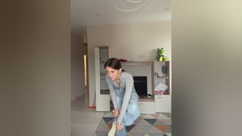 Video of a woman vacuuming a living room with beige walls, a green plant, and a patterned rug. She wears a grey top and blue overalls.