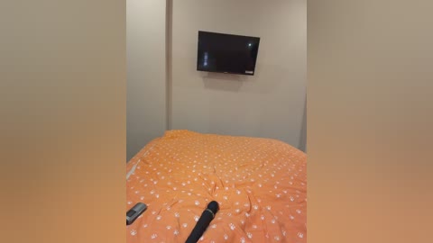 A video of a simple bedroom with an orange bedspread featuring white polka dots. A flat-screen TV mounted on the beige wall hangs above the bed, and a remote control lies on the bed.
