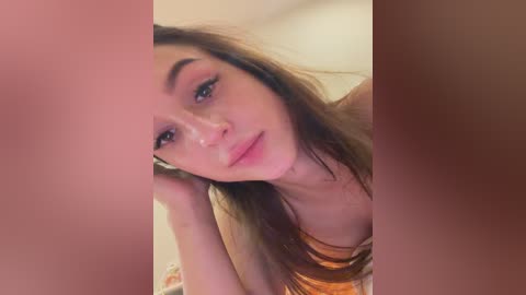 A close-up video of a young woman with fair skin, brown hair, and light makeup, resting her head on her hand, wearing a sleeveless, orange top. The background is blurred.