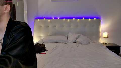 A video of a modern bedroom with a plush, tufted headboard, lit by blue LED lights. A man in a black jacket stands beside the bed, which has white pillows and a red object on the nightstand.