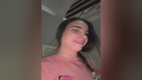 Video of a young woman with long dark hair, wearing a pink T-shirt, smiling, standing indoors, with blurred background featuring air conditioning and a gray wall.