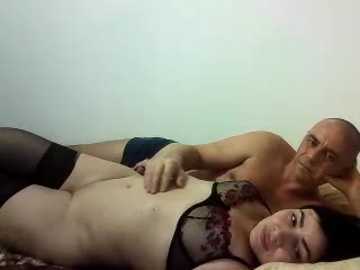 Video of a bald, muscular man in black shorts, lying on a bed with a fair-skinned woman in black lingerie, her dark hair splayed on a pillow, both gazing at each other.