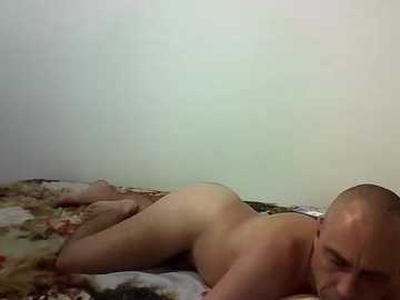 Video of a bald, fair-skinned man lying naked on a floral-patterned bed, with his face turned away, in a dimly lit room with plain white walls.